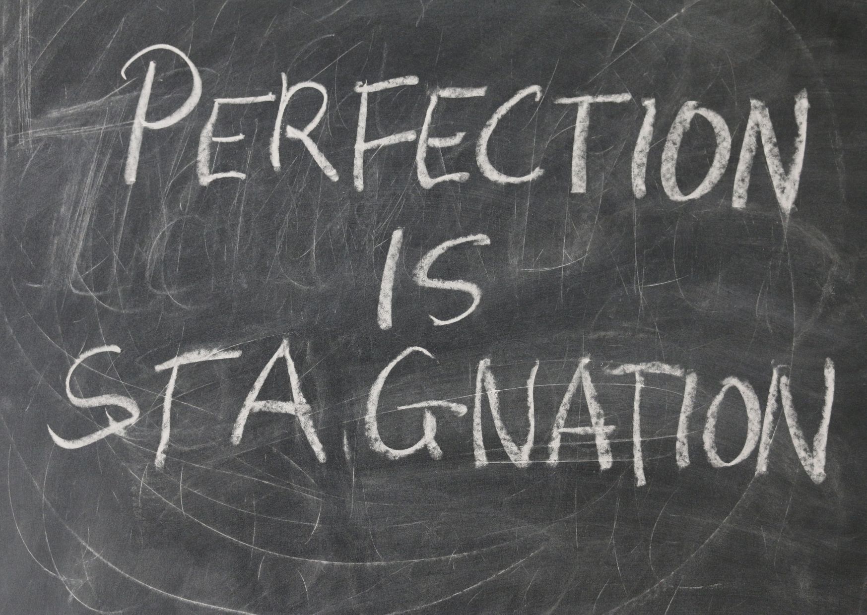 Na šolski tabli je s kredo napisano ‘Perfection is stagnation’, kar poudarja idejo, da popolnost lahko zavira napredek.