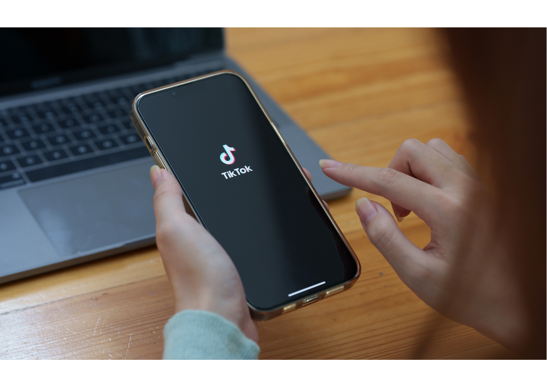 Oseba drži pametni telefon z odprto aplikacijo TikTok, v ozadju je prenosni računalnik na mizi.