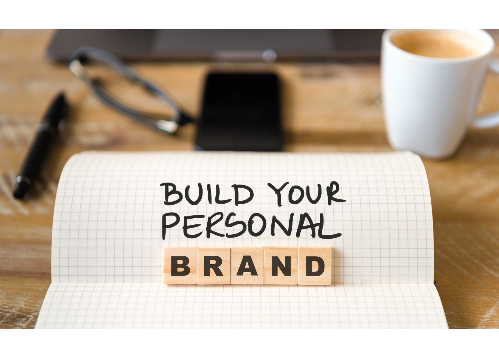 Zvezek na mizi z napisom ‘Build your personal brand’, obdan s pisalom, očali, telefonom in skodelico kave, kar simbolizira razvoj osebne blagovne znamke in profesionalne identitete
